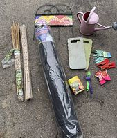 Full view of garden fabric roll in plastic wrapper with bundles of wooden stakes, metal plant hanger, watering can, garden knee pads, gloves, seed packet, and small hand tools.
