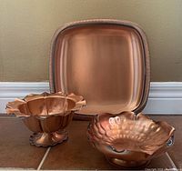 Photo of three mid-century hand hammered copper serving pieces: a square tray and two decorative bowls with hammered textures and ridged or ruffled designs.