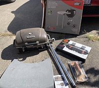 Two portable grills including a brand new Weber hibachi grill with gray carrying case, a used foldable grill with metal legs, grilling tools and parts on pavement with boxed Weber work table in background