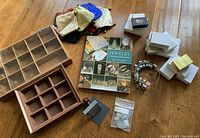 Jewelry maker supplies arranged on wooden floor including Tim McCright's manual, wooden storage boxes with compartments, silk pouches, jewelry boxes, metal ring sizer, stamp handle with lino piece, metal price tags, and silver-tone jewelry.