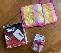 Overall lot showing all items: Black & Decker hand mixer box, two Trudeau sprinkle jars with pink lids, and two pastel quilted potholders.