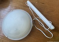 Large flushmount light dome and white plug-in tube light laid on wooden floor, showing the front view of both items.