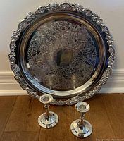 Silver plated round serving tray with intricate floral edge and engraved center leaning against wall with two candlesticks in front