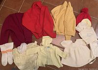 Overall lot layout showing an assortment of vintage knit baby clothes including sweaters, cardigans, dresses, hats, socks and booties in various colors primarily cream, yellow, maroon, red, and white.