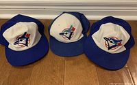 Photo of three vintage Toronto Blue Jays baseball caps arranged side by side on a wooden floor.