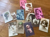 Group of vintage postcards and sheet of buttons laid out on wood floor. Postcards primarily depict women in vintage fashion from late 1800s to early 1900s with some tinted colors and sepia tones.