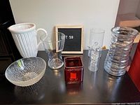 Photo showing six assorted glass and ceramic items including pitcher, bowl, several vases, and glass pipe arranged on a black surface.