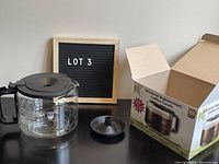 Clear glass coffee carafe with black plastic lid and handle displayed next to its original packaging box and letter board indicating lot number