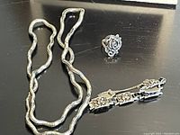 Photo of three silver-coloured jewelry items: a wavy chain necklace, a floral design ring, and a floral link bracelet with a clasp, displayed together on a black surface.