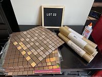 Nine metallic copper-colored peel and stick mosaic tile sheets and four rolls of gold glitter fabric displayed on a table alongside a bottle of glue and LOT 22 sign