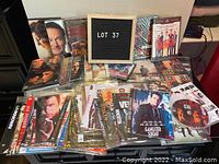 Image shows a collection of assorted DVDs arranged on table, some in plastic sleeves and some in cases, with a letter board labeled 'LOT 37'
