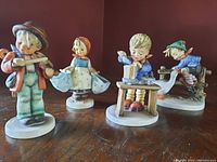 Four Hummel figurines displayed on a wooden surface showing a boy playing fiddle, girl holding pail, boy at table cooking, child and goose.