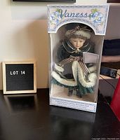 Full view of Vanessa porcelain doll inside original display box labeled 'Vanessa' with floral design. Box shows wear with opening at display area.