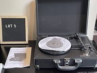 Black suitcase-style vintage Replica turntable open with a vinyl record on platter, manual booklet on the left, 'LOT 5' sign in the background.