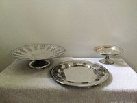 Three vintage silver plate serving dishes on a white cloth; one large pedestal dish with pierced edge, one flat plate, and one smaller pedestal dish