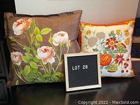 Two floral silk pillows displayed on black surface, one large brown with pink roses and one smaller white with colorful flowers and fruits