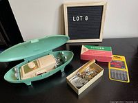 Vintage Singer button holer attachment in light green oval case with cover open, showing the attachment inside, accompanied by a box of assorted sewing machine attachments and a pack of Singer needles.