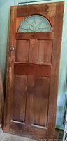 Full view of solid wooden door showing four panels, wood grain, and arched leaded glass transom window at top. Door has a round metal knob on the left side and some visible wood damage and discoloration.