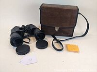 Black Bushnell binoculars with neck strap and lens caps removed showing lenses, next to brown carrying case and yellow lens cloth.