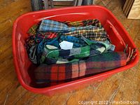 Red plastic container with folded plaid wool items inside including blankets and garments.