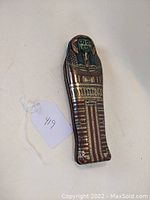 Front view of lithographed tin sarcophagus figurine showing detailed painted green face and sarcophagus decorations.