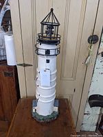 Full view of the white ceramic/resin lighthouse solar light with black metal top railing and pointed spire, standing on wooden surface against paneled wood background.