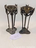 Pair of bronze font-style basins on three-legged tripod stands showing front and side views.