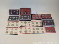 Wide view of multiple United States Mint Proof and Mint Sets from 1974 to 1990, all featuring clad coins. Sets are sealed or in protective holders, some missing packaging.
