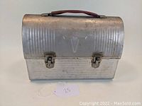 Front view of steel dome top lunch box showing the rounded lid with vertical ribbing and a faint 'V' embossed on the front center. Visible metal latches and brown handle.
