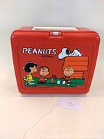 Front view of red Peanuts plastic lunch box illustrating several Peanuts characters including Lucy, Linus, Snoopy, Woodstock, and Charlie Brown around Snoopy's doghouse, with the brand 'Peanuts by Schulz' prominently displayed.