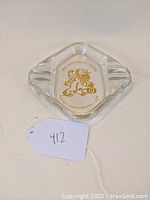 Clear glass square ashtray with the Ritz-Carlton gold lion logo and lettering, side views showing cigarette rests