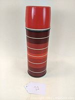 Front view of a tall vintage thermos with multi-toned red horizontal stripes and solid red plastic lid.