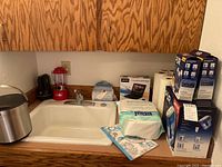 Overall lot items arranged on kitchen counter around a white sink: ice bucket, flashlight, lantern, Epsom salt package, sleep sack, portable DVD player box, PUR water filter pitcher and replacements, paper towels.