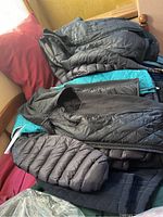 Multiple women's coats piled on a couch, showing various styles and colors including black and blue, with quilted and puffer designs.