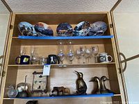 Photo of wooden shelf displaying collectible wildlife decorative plates on top shelf, various glassware including decanter and mugs on middle shelf, and metal, wooden, and glass animal figurines on bottom shelf.