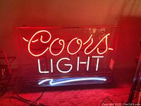 Front view of neon sign with 'Coors' in red, 'LIGHT' in white, and blue wave underneath, lit up on carpeted floor against red wall.