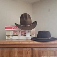 Two cowboy hats displayed on a wooden cabinet, one sitting on a Bailey shoe box, one flat next to it.