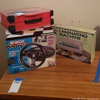 Photo showing the red tool case, NASCAR Thrustmaster racing wheel box, and Royal Sovereign laminating machine box on a wooden surface.