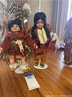 Two Native American collectible ceramic dolls approximately 13 inches tall, shown standing on white doll stands on wood surface with cultural artifacts in the background.