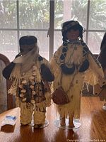 Two Native American collectible ceramic dolls seen from front in natural light with detailed traditional clothing including fringes, beads, baskets, and headbands.