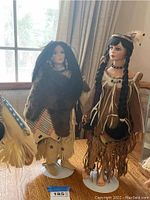 Two Native American ceramic collectible dolls with stands displayed on table in front of window.