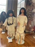 Front angled view of two Native American collectible ceramic dolls wearing traditional fringed dresses with beadwork, standing on round white stands.