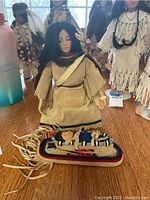 Two Native American ceramic collectible dolls displayed on a wooden surface with other similar collectible items in the background.