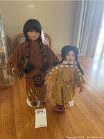 Two native American collectible ceramic dolls standing on individual white stands, placed on a table in natural lighting. Both dolls have braided hair and traditional clothing.