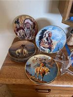 Six Native American collectible decorative plates with detailed painted scenes of Native Americans and horses, many with feather headdresses, all with gold trim.