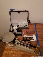 Ohaus Triple Beam Balance Scale with metal pan, two vintage cameras, silver carrying case, and wooden box of weights on wooden surface against plain wall.