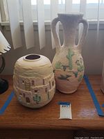 Two Southwestern style pottery vases on wood table under window. One rounded with pueblo adobe motifs, the other tall with handles and desert cactus and animals.