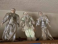 Three vintage dolls on shelf, dressed in silver and cream colors, visible feather and lace details