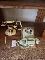 Two vintage rotary dial telephones displayed on a wooden shelf: one ivory candlestick-style and one beige desk rotary phone.