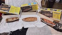 Three wooden model boats (Whitehall, Dinghy, Peapod) displayed with their boxes behind, showing product packaging and models out of box on a cloth surface.